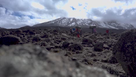 Toma-Estática-De-Personas-Que-Llevan-Equipaje-En-La-Cabeza,-Caminando-Lentamente,-Con-El-Monte-Kilimanjaro-Al-Fondo