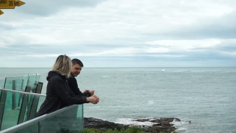 Young-attractive-couple-looking-at-ocean-by-Stirling-Point-sign-in-Bluff,-New-Zealand