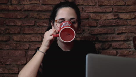 Close-up-shot-of-Young-beautiful-brunette-wearing-glasses-watching-the-computer-and-drinking-coffee-of-a-cup-with-the-inscription-"Just-one-more-episode