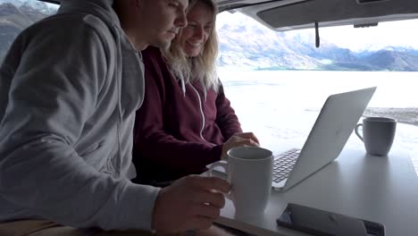 SLOWMO---Young-attractive-couple-sitting-in-back-of-motorhome-interacting-with-laptop-by-Lake-Wakatipu,-Queenstown,-New-Zealand