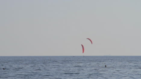 Two-kite-surfers-sail-by-a-catamaran,-near-the-beach