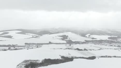 Aerial-right-pan-through-snowed-in-hill-landscape-with-town-and-mountains-in-background