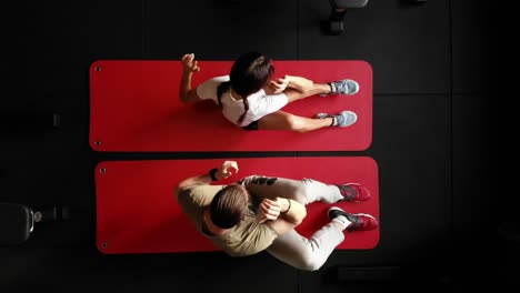 Top-down-drone-shot-of-sporty-woman-exercising-in-gym-on-mat-with-personal-trainer