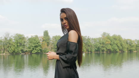 Young-Russian-lady-wearing-a-black-dress,-stands-in-front-of-a-lake,-surrounded-by-trees