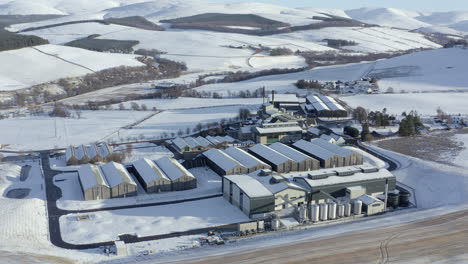Aerial-view-of-the-Glenlivet-whisky-distillery-surrounded-by-snow-in-the-late-afternoon-sun-on-a-winters-day,-Moray,-Scotland---advancing-shot