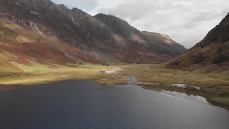 Slowly-pulling-back-along-a-dark-lake-to-reveal-a-Scottish-highland-valley-in-Glencoe,-Scotland