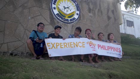 Junge-Leute-Lächeln-Und-Winken,-Während-Sie-Ein-Weißes-Banner-Mit-Der-Aufschrift-„experience-Philippines“-Schwenken