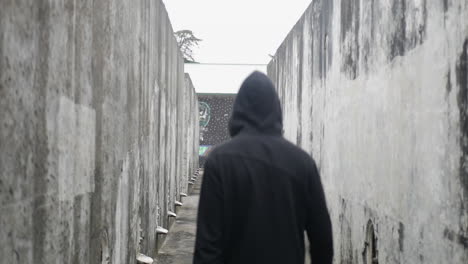 Man-wearing-a-black-hoodie-looking-around-him-while-walking-furtively-in-an-abandoned-structure-with-graffiti,-in-a-cloudy-day