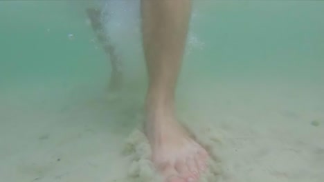 Slow-motion-underwater-view-of-feet-walking-on-sandy-ocean-floor-in-shallow,-clear-ocean-water