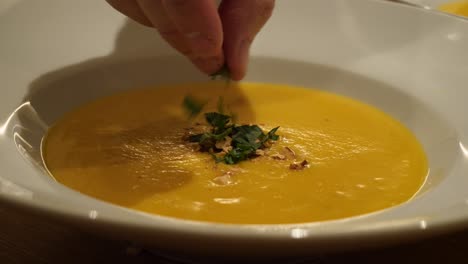 A-white-male-hand-is-sprinkling-herbs-on-a-yellow-soup-in-a-white-plate-in-slow-motion