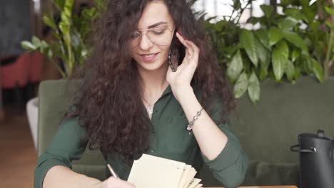 Junge-Geschäftsfrau-Spricht-Auf-Ihrem-Telefon-Und-Schreibt-In-Ihr-Notizbuch,-Während-Sie-In-Einem-Café-Arbeitet