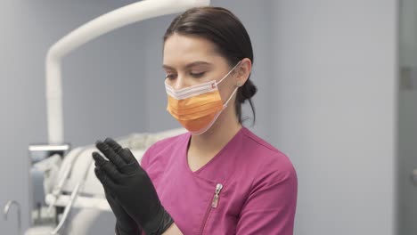 Female-Dentist-in-Dental-Clinic-Office-Preparing-for-Exam-and-Treatment-Wearing-Black-Gloves-and-Correcting-Mouth-Mask-Position-While-Looking-at-the-Camera