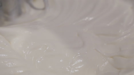 Close-up-macro-view-of-metal-mixer-stirring-and-blending-thick,-white-icing-in-large-bowl-in-slow-motion