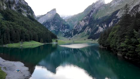 Aerial-flyover-low-over-the-water-of-lake-Seealpsee-in-Appenzell,-Switzerland-with-a-reflection-of-the-Alpstein-peaks-on-the-lake's-still-surface