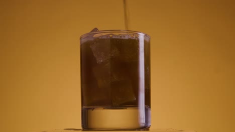 Static-shot-of-glass-filled-with-ice-cubes-against-golden-yellow-background-with-orange-light-setting-into-which-cola-is-filled