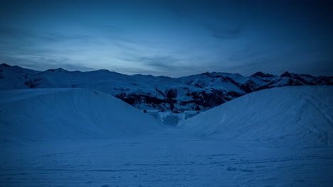 Sunset-time-lapse-from-within-half-pipe-at-ski-resort---snowy-mountains-and-snow-groomers-preparing-slopes-with-lights-on