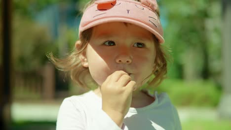 Face-of-blond-two-year-old-girl-sucking-and-slowly-eating-white-vitamin-pill-while-standing-in-a-park-and-looking-at-camera-daytime
