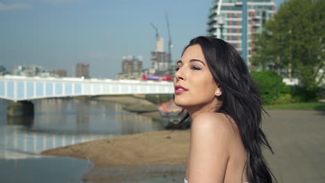 Slow-Motion-of-a-Beautiful-latina-woman-on-holiday-leaning-against-the-railing,-looking-at-the-river-Thames-in-London-and-waving-with-her-hand,-smiling-and-wandering