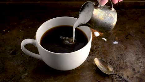 Tasse-Kaffee-Mit-Dicker-Sahne,-Die-Langsam-Aus-Einem-Kleinen-Metallkännchen-In-Dunkler-Umgebung-Eingegossen-Wird