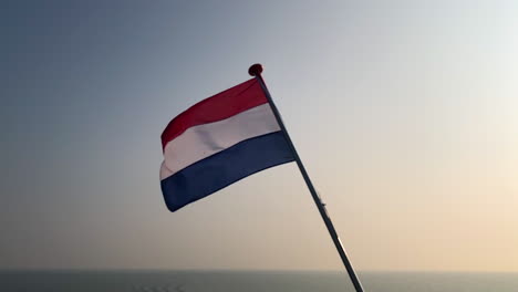 Slow-motion-of-the-dutch-flag-of-the-Netherlands-in-the-wind-at-sunset-over-a-background-of-moving-water-from-the-north-sea