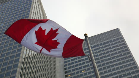 Low-angle-shot,-low-angle-view-of-canada-flag-waving-in-wind,-and-skyscraper-as-background