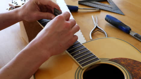 Hands-of-a-luthier-craftsman-measuring-and-leveling-an-acoustic-guitar-neck-fretboard-on-a-wood-workshop-bench-with-lutherie-tools