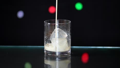 Pouring-almond-milk-into-a-glass-filled-with-ice,-sitting-on-a-glass-surface-with-a-black-background-with-fairy-lights