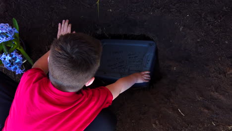 A-young-boy-placing-the-plastic-case-of-the-body-of-a-dead-family-pet-in-a-recently-dug-hole-in-a-family-garden-to-bury-them-there-and-have-their-grave-in-the-garden