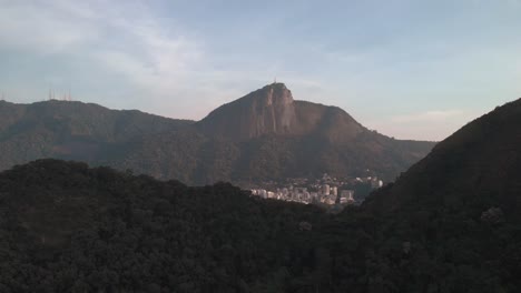 Sunrise-backwards-aerial-view-of-the-Corcovado-mountain-in-Rio-de-Janeiro-revealing-a-mountain-range-in-the-foreground-elsewhere-in-the-city-with-high-rise-residential-buildings-in-front