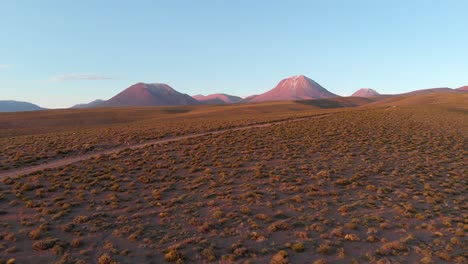 Toma-Cinematográfica-Aérea-De-Un-Camino-De-Tierra-Con-Volcanes-Y-Montañas-Nevadas-En-El-Fondo-En-El-Desierto-De-Atacama,-Chile,-Sudamérica