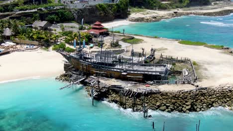 Old-wooden-ship-moored-on-stones-in-tropical-beach-of-Bali,-aerial-orbit-view