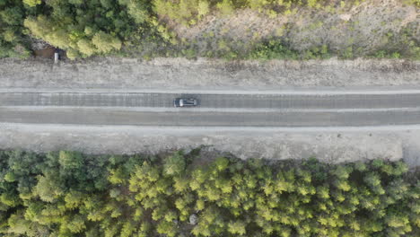 4k-Aerial-top-view-over-a-car-driving-on-a-dirt-country-forest-road-with-green-trees-of-dense-forests-growing-both-sides