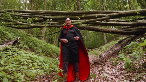 Portrait-Of-A-Man-Dressed-Up-In-Black-And-Red-Cape-Taking-Off-His-Hood-While-Walking-On-A-Trail-In-The-Forest