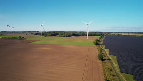 Panning-aerial-view-passing-across-sustainable-solar-panel-array-and-wind-turbines-on-rural-farmland