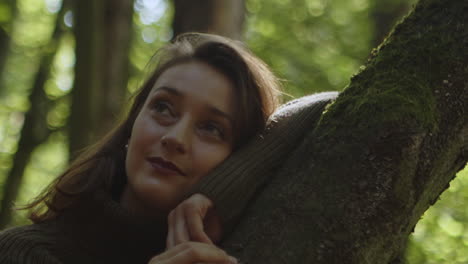 Beautiful-brunette-with-dreamy-eyes-is-leaning-against-a-tree-trunk-surrounded-by-forest-nature
