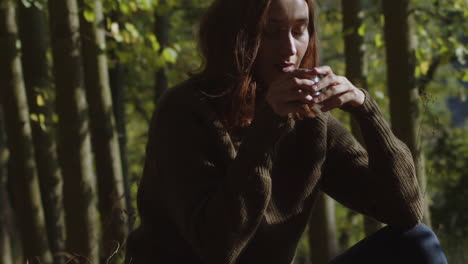 A-young-woman-sitting-in-the-grass-in-the-middle-of-the-forest-enjoying-a-warm-cup-of-coffee-from-a-thermos