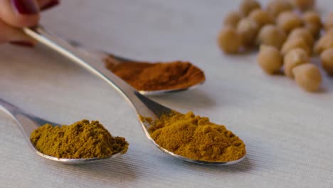 A-spoonful-of-curry-powder-is-placed-on-a-table-next-to-two-other-spoons-with-turmeric-powder-and-saffron-powder,-chickpeas-can-be-seen-in-the-background