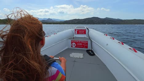 Back-view-of-long-hair-redhead-child-with-sunglasses-on-motorboat-sailing-over-north-Corsica-sea-water-for-tour-toward-Saleccia-famous-beach-1
