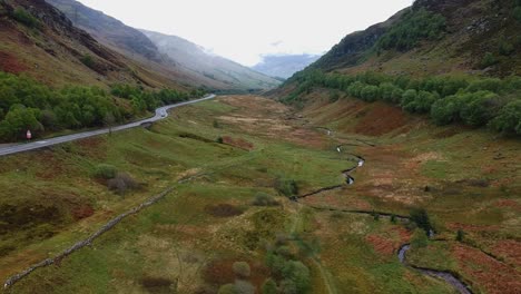Cinematic-drone-shot-of-scottish-highland-valley-with-car-passing-by-on-windind-road-in-background