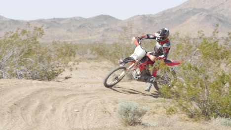 SLOW-MOTION:-A-dirt-biker-rides-his-red-motorcycle-through-the-desert-and-whips-around-a-berm