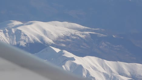 Zoomed-out-view-of-snow-capped-mountains-while-looking-out-plane-window-and-flying-over-europe-with-jet-wing-in-frame