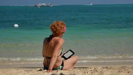 Eine-Aufnahme-Von-Hinten-Einer-Rothaarigen-Frau,-Die-Am-Strand-Ein-E-Book-Liest-Und-Einen-Bikini-Trägt