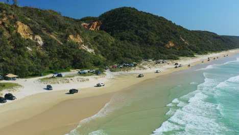 Aerial-Convoy-Of-Cars-Driving-Along-Beach-Camp-Sites-Beside-Blue-Ocean-Waves-In-Queensland,-4K-Drone