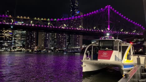 CityHopper-docked-at-howard-smith-wharves-ferry-terminal-waiting-for-its-schedule-ride-at-night-with-iconic-story-bridge-lit-up-in-purple,-reflected-on-wavy-river-water-and-urban-downtown-cityscape