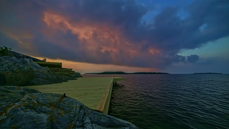 Colorful-sunset-cloudscape-over-a-lake-or-fjord-with-a-wooden-dock-in-the-rocky-foreground---sliding-time-lapse