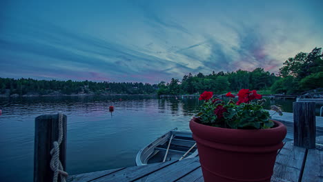 Overview-shot-of-a-red-potted-flower-on-wooden-pier-along-rocky-slope-beside-a-calm-lake-on-in-timelapse