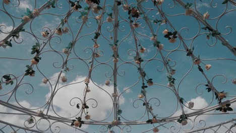 Nahaufnahme-Eines-Zeremoniellen-Metallbogens,-Geschmückt-Mit-Weißen-Und-Rosa-Rosen,-über-Einem-Wunderschönen-Blauen-Sommerhimmel-Und-Weißen-Wolken