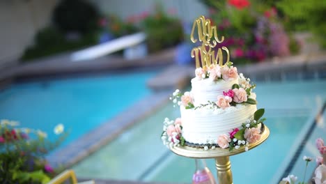 Shot-of-a-beautifully-decorated-floral-wedding-cake-sitting-on-a-pedestal-right-next-to-a-pool-during-a-summer-wedding-reception