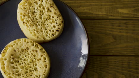 Top-down-shot-of-a-male-placing-some-tasty-and-delicious-crumpets-on-a-blue-stone-plate-on-a-wooden-background,-buttering-them-then-taking-a-bite-and-placing-back-down