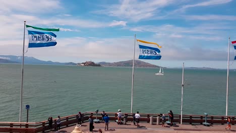 Shot-of-flags-from-Pier-39-waving-in-strong-wind-at-the-end-of-Pier-39-with-Alcatraz-Island-on-a-summer-day-in-San-Francisco,-California,-USA